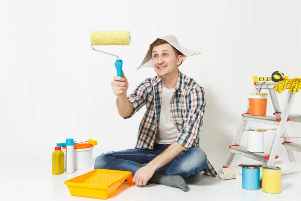 Ο άνθρωπος με εφημερίδα καπέλο που κάθονται στο πάτωμα, χρησιμοποιώντας ρολό βαφής, όργανα για ανακαίνιση Διαμέρισμα Δωμάτιο απομονωθεί σε λευκό φόντο. Ταπετσαρία, κόλλημα αξεσουάρ, εργαλεία βαφής. Επισκευή concept home. — Φωτογραφία Αρχείου