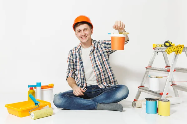 Homem de capacete de proteção laranja sentado no chão com lata de tinta, instrumentos para renovação apartamento isolado no fundo branco. Papel de parede, acessórios de colagem, ferramentas de pintura. Reparação home concept . — Fotografia de Stock