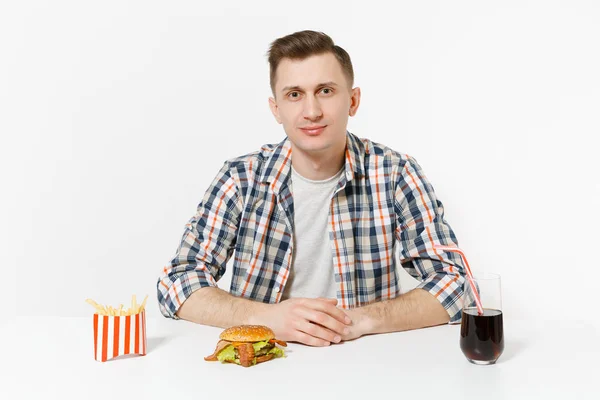 Bonito jovem de camisa sentado à mesa com hambúrguer, batatas fritas, cola em vidro isolado sobre fundo branco. Nutrição adequada ou fast food clássico americano. Área de publicidade com espaço de cópia . — Fotografia de Stock