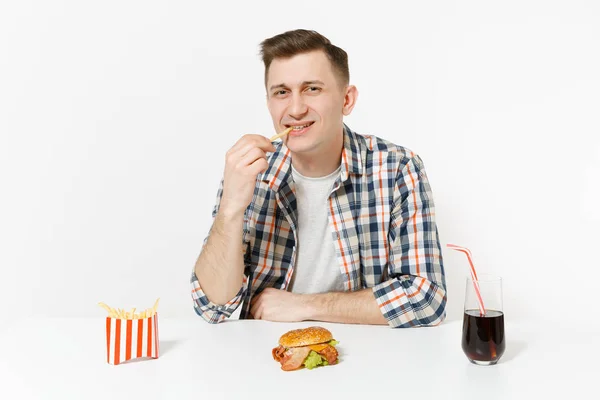 Szórakoztató éhes fiatal ember eszik a francia krumpli, ül az asztal burger, cola pohár elszigetelt fehér background. Megfelelő táplálkozás, vagy a klasszikus amerikai gyorsétterem. Hirdetés terület másol hely. — Stock Fotó