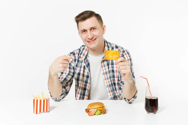 Knappe jonge man zit aan tafel met creditcard, burger, frietjes, cola in het glas geïsoleerd op een witte achtergrond. Goede voeding of Amerikaanse klassieke fastfood. Gebied met kopie ruimte reclame — Stockfoto