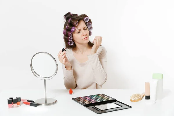 Mujer divertida con rizadores pintando uñas con esmalte de uñas rojo, sentada a la mesa aplicando maquillaje con cosméticos decorativos faciales establecidos aislados sobre fondo blanco. Belleza mujer moda estilo de vida concepto . — Foto de Stock