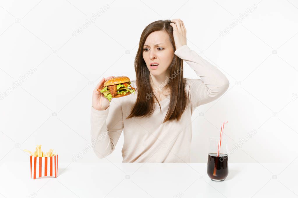 Shocked young woman sitting at table with burger, french fries, cola in glass bottle isolated on white background. Proper nutrition or American classic fast food. Advertising area with copy space.