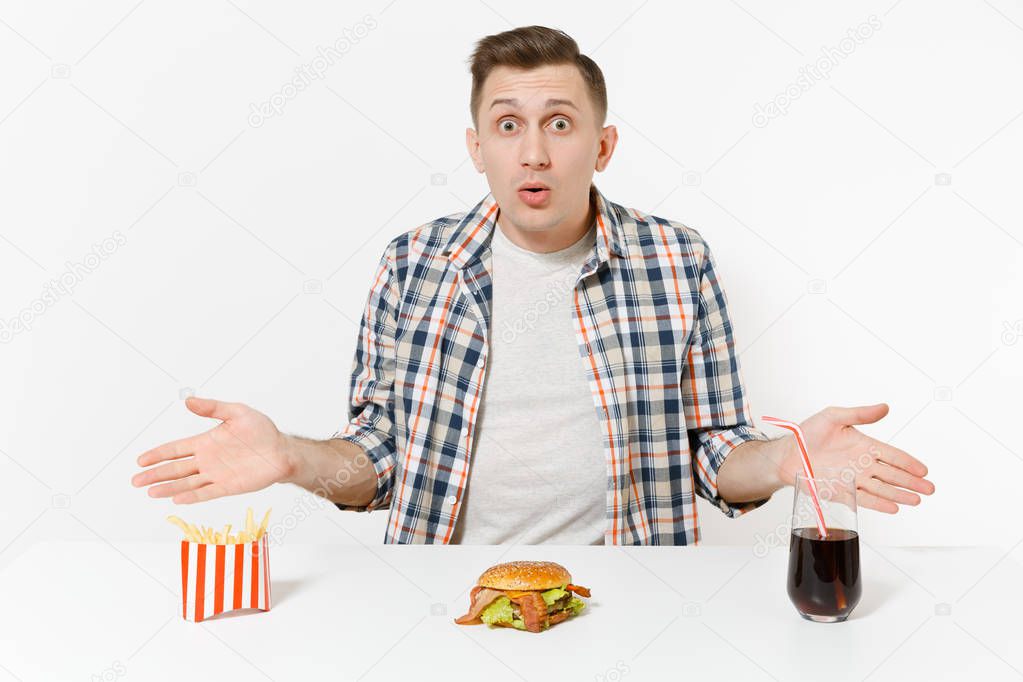 Shocked young man spreading hands, sitting at table with burger, french fries, cola in glass isolated on white background. Proper nutrition or American classic fast food. Advertising area, copy space.