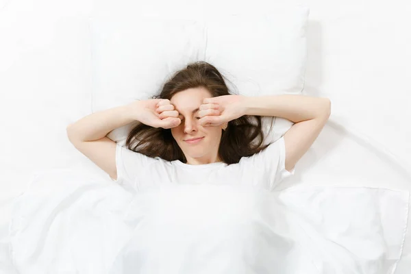 Top view of head of happy brunette young woman lying in bed with white sheet, pillow, blanket. Smiling female stretching out in bed, spending time in room. Rest, relax, good mood concept. Copy space. — Stock Photo, Image