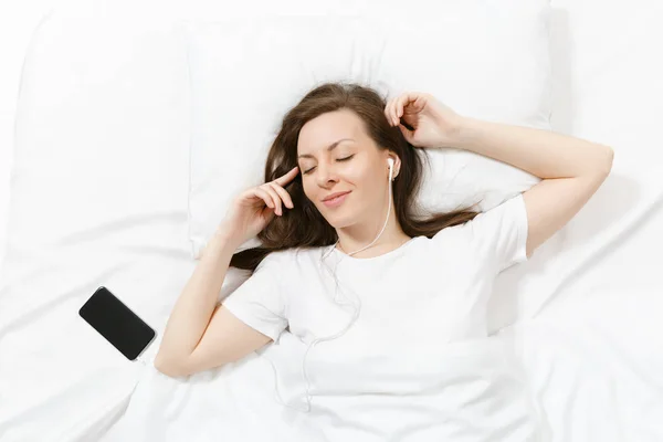 Üstten Görünüm mutlu genç kadının beyaz çarşaf, yastık, battaniye, dinle müzik cep telefonu kulaklık ile yatakta yalan. Odada vakit sakin güzel kadın. Dinlenmek, dinlenmek, iyi ruh kavramı.