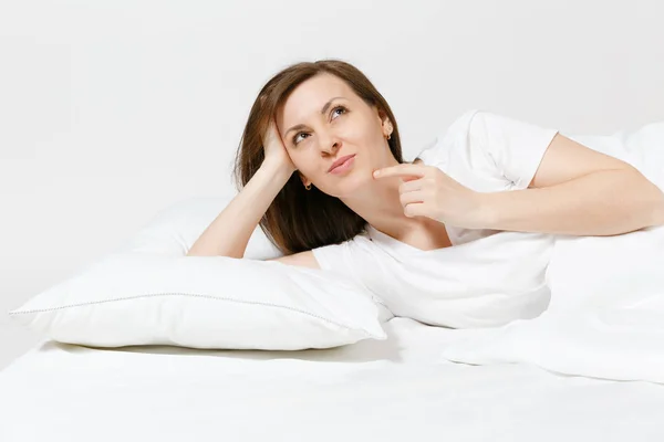 Calm young brunette woman lying in bed with white sheet, pillow, blanket on white background. Dreamful beauty female spending time in room. Rest, relax, good mood concept. Copy space for advertisement — Stock Photo, Image