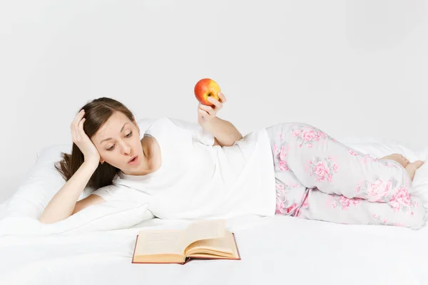 Mujer joven sentada en la cama con sábana blanca, almohada, manta sobre fondo blanco. Mujer pasando tiempo en la habitación, leyendo libros, comiendo manzana, disfrutando del descanso. Relájate, concepto de buen humor. Lugar para el texto . — Foto de Stock