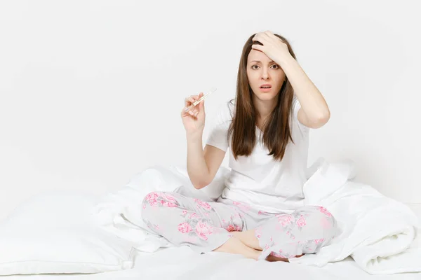 Mujer triste cansada joven enferma en pijama sentada en la cama, sosteniendo termómetro clínico con alta temperatura de fiebre aislada sobre fondo blanco. Mujer sintiéndose mal, pasando tiempo en casa. Área publicitaria — Foto de Stock