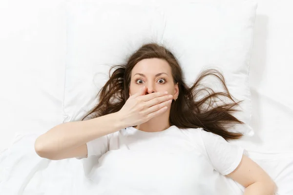 Top view of head of tired brunette young woman lying in bed with white sheet, pillow, blanket. Shocked female cover mouth with hand, spending time in room. Rest, relax, good mood concept. Copy space.