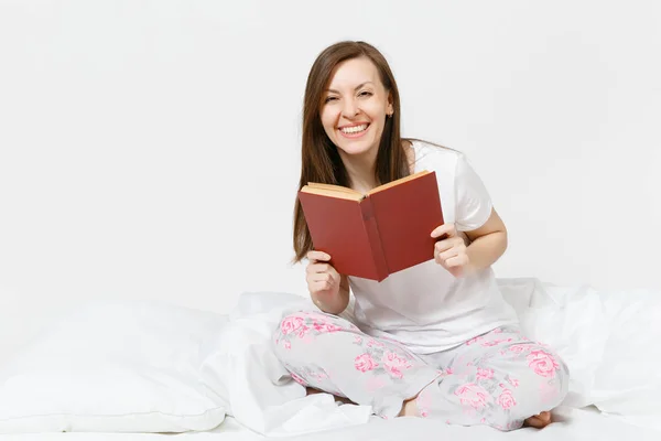 Jovem mulher feliz sentada na cama com lençol branco, travesseiro, cobertor sobre fundo branco. Beleza feminina passar o tempo no quarto, ler livro, desfrutar de descanso. Relaxa, conceito de bom humor. Lugar para texto . — Fotografia de Stock