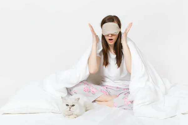 Lugn ung kvinna sitter i sängen med sovmask på ögon isolerad på vit bakgrund. Skönhet kvinna umgås i rum med vita söta Persiska silver chinchilla katt. Vila, koppla av, gott humör koncept. — Stockfoto