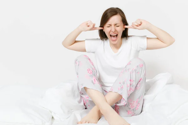 Stressed crazy mad young screaming woman sitting in bed with white sheet, pillow, blanket. Shocked female cover ears with hands, spending time in room. Bad mood, stress, problem concept. Copy space. — Stock Photo, Image