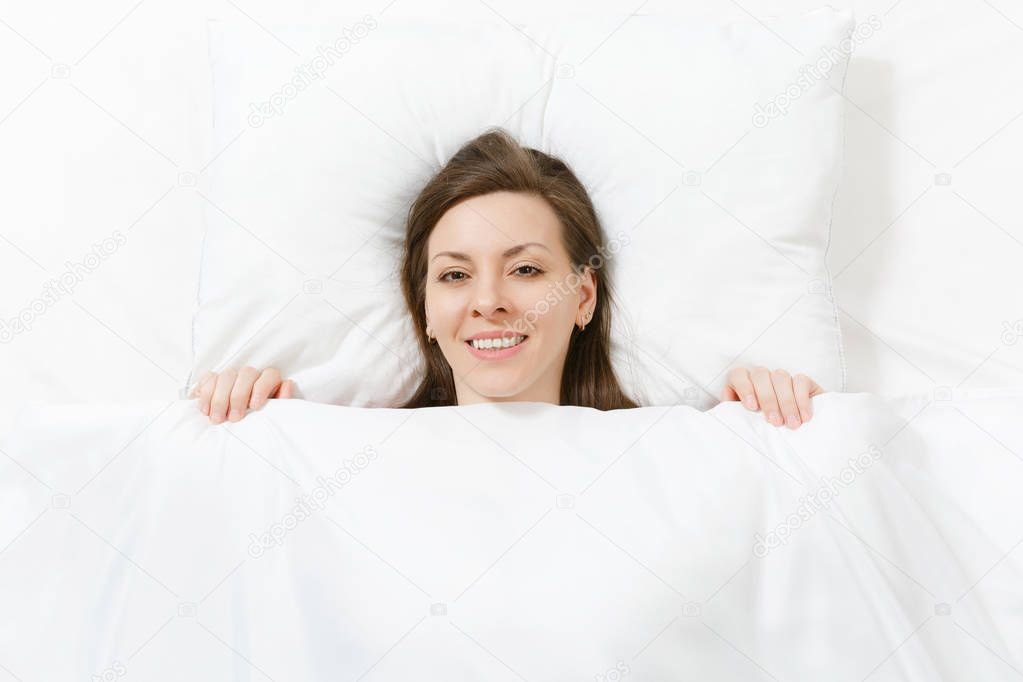 Top view of head of happy brunette young woman lying in bed with white sheet, pillow, blanket. Smiling pretty female spending time in room. Rest, relax, good mood concept. Copy space for advertisement