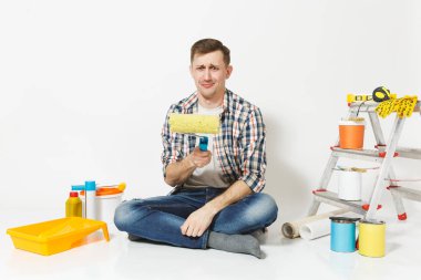 Dissatisfied upset man sitting on floor with paint roller, instruments for renovation apartment room isolated on white background. Wallpaper, gluing accessories, painting tools. Repair home concept. clipart