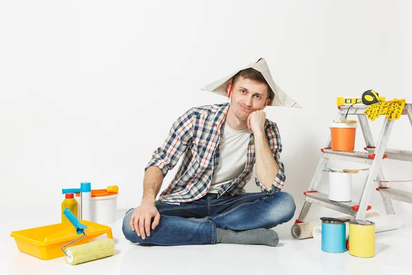 Homem de chapéu de jornal com lápis atrás da orelha sentado no chão com instrumentos para renovação apartamento isolado no fundo branco. Papel de parede, acessórios de colagem, ferramentas de pintura. Conceito de reparação . — Fotografia de Stock