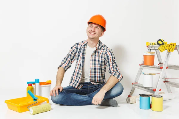 Homem sonhador em capacete de proteção laranja sentado no chão com instrumentos para renovação apartamento quarto isolado no fundo branco. Papel de parede, acessórios de colagem, ferramentas de pintura. Reparação home concept . — Fotografia de Stock