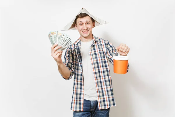 Jeune homme souriant au chapeau de journal tenant un paquet de dollars, de l'argent comptant et un seau de peinture vide avec un espace de copie isolé sur fond blanc. Instruments pour salle de rénovation. Réparation concept de maison . — Photo