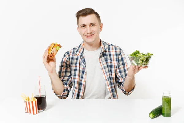 El hombre se sienta a la mesa con batidos de desintoxicación verde, ensalada en tazón de vidrio, pepino, hamburguesa, papas fritas, cola en vidrio aislado sobre fondo blanco. Nutrición adecuada, estilo de vida saludable, comida rápida, concepto de elección . — Foto de Stock