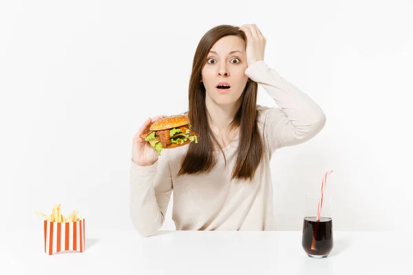 Giovane donna scioccata seduta a tavola con hamburger, patatine fritte, cola in bottiglia di vetro isolata su fondo bianco. Una corretta alimentazione o un fast food classico americano. Area pubblicitaria con spazio di copia . — Foto Stock