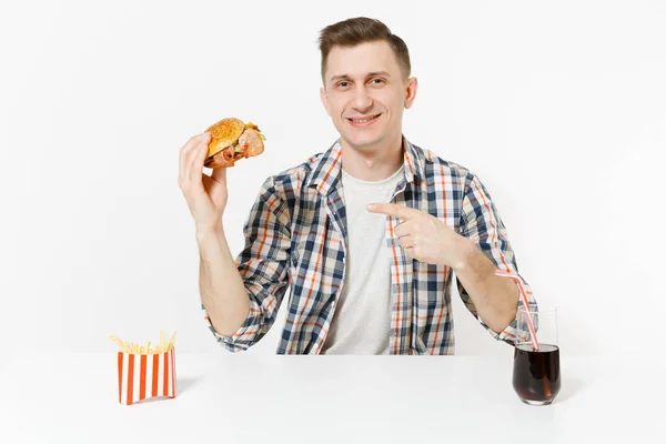 Bonito jovem sorridente sentado à mesa com hambúrguer, batatas fritas, cola em vidro isolado no fundo branco. Nutrição adequada ou fast food clássico americano. Área de publicidade com espaço de cópia . — Fotografia de Stock