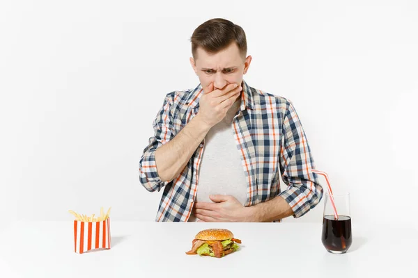 Homem doença colocar as mãos na dor abdominal, dor de estômago à mesa com hambúrguer, batatas fritas, cola em vidro isolado no fundo branco. Nutrição adequada ou fast food clássico americano. Área com espaço de cópia — Fotografia de Stock