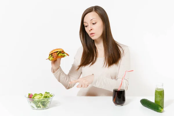Woman at table with green detox smoothies, salad in glass bowl, cucumber, burger, cola in bottle isolated on white background. Proper nutrition, healthy lifestyle, classic fast food, dieting concept. Stock Photo