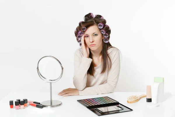 Mujer triste cansada con rizadores en el pelo sentado en la mesa aplicando maquillaje con cosméticos decorativos faciales establecidos aislados sobre fondo blanco. Belleza concepto de estilo de vida femenino. Área con espacio de copia . — Foto de Stock