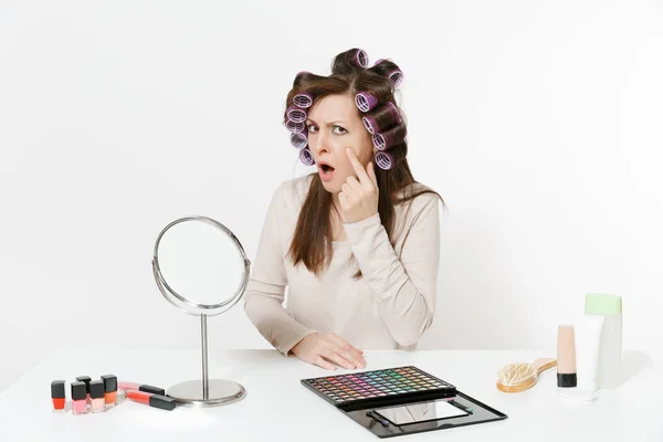 Mujer insatisfecha con rizadores en el pelo sentado en la mesa aplicando maquillaje con cosméticos decorativos faciales establecidos aislados sobre fondo blanco. Belleza concepto de estilo de vida femenino. Área para copiar espacio . — Foto de Stock