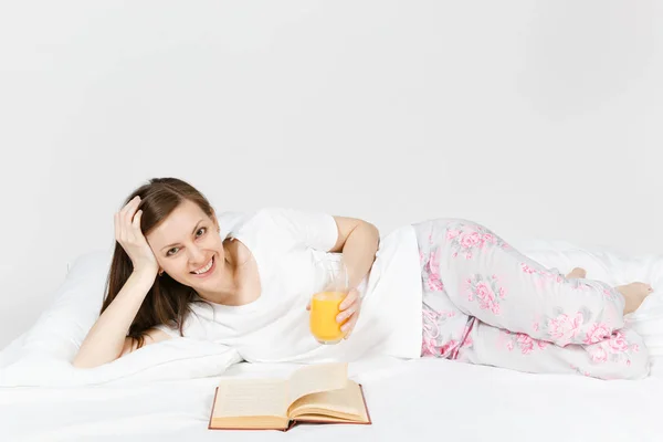 Mujer joven sentada en la cama con sábana blanca, almohada, manta sobre fondo blanco. Mujer pasando tiempo en la habitación, leyendo libros, comiendo manzana, disfrutando del descanso. Relájate, concepto de buen humor. Lugar para el texto . — Foto de Stock