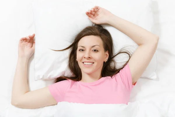 Top view of happy brunette young woman lying in bed with white sheet, pillow, blanket, hands under head. Smiling female spending time in room. Rest, relax, good mood concept. Copy space advertisement.