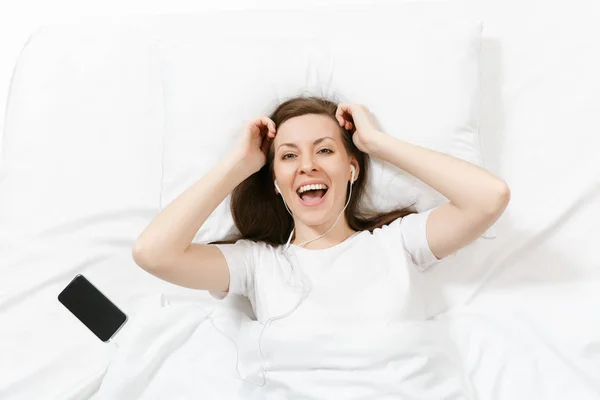 Top view of happy young woman lying in bed with white sheet, pillow, blanket, listen music from earphones in mobile phone. Calm beautiful female spending time in room. Rest, relax, good mood concept. — Stock Photo, Image
