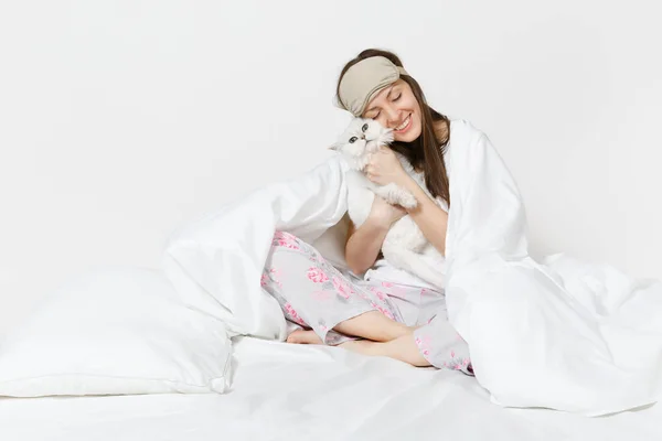 Lugn ung kvinna sitter i sängen med sovmask på ögon isolerad på vit bakgrund. Skönhet kvinna umgås i rum med vita söta Persiska silver chinchilla katt. Vila, koppla av, gott humör koncept. — Stockfoto
