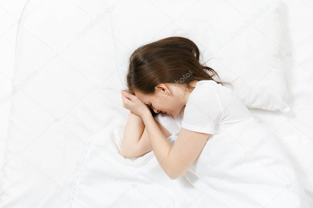Top view of tired stressed crying young brunette woman lying in bed with white sheet, pillow, blanket. Shocked frustrated sad upset female lies on her side. Rest, relax, bad mood concept. Copy space.
