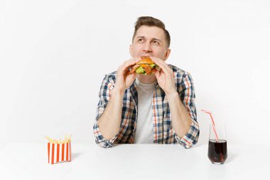 Hamburger yeme, tablo ile kızarmış patates, kola cam beyaz arka plan üzerinde izole oturup eğlenceli aç genç adam. Doğru beslenme veya Amerikan Klasik fast food. Alan kopya alanı ile reklam.