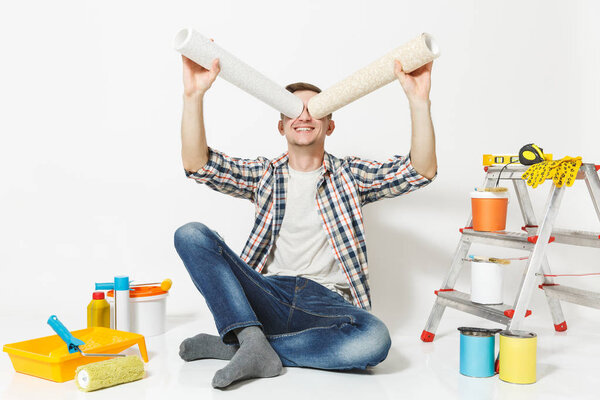 Fun male look through rolls of wallpaper as in binoculars, sit on floor, instruments for renovation apartment isolated on white background. Gluing accessories, painting tools. Repair home concept.