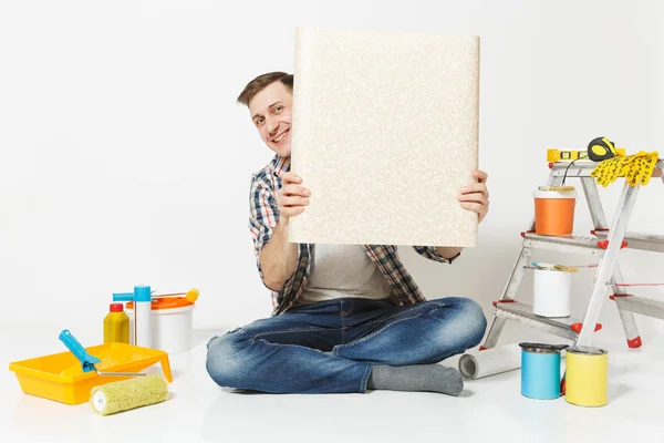 Hombre guapo sentado en el suelo con rollo desplegado de instrumentos de papel pintado para la renovación habitación del apartamento aislado sobre fondo blanco. Pegado de accesorios herramientas de pintura. Reparar concepto de casa. Copiar espacio — Foto de Stock