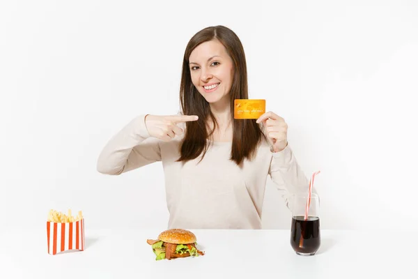 Mujer feliz sentada en la mesa con tarjeta de crédito, hamburguesa, papas fritas, cola en botella de vidrio aislado sobre fondo blanco. Nutrición adecuada o comida rápida clásica americana. Área de publicidad con espacio de copia —  Fotos de Stock