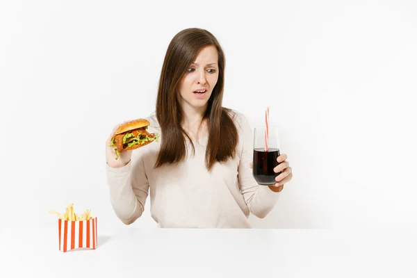 Dissatisfied woman sitting at table with burger, french fries, cola in glass bottle isolated on white background. Proper nutrition or American classic fast food. Advertising area with copy space. — Stock Photo, Image