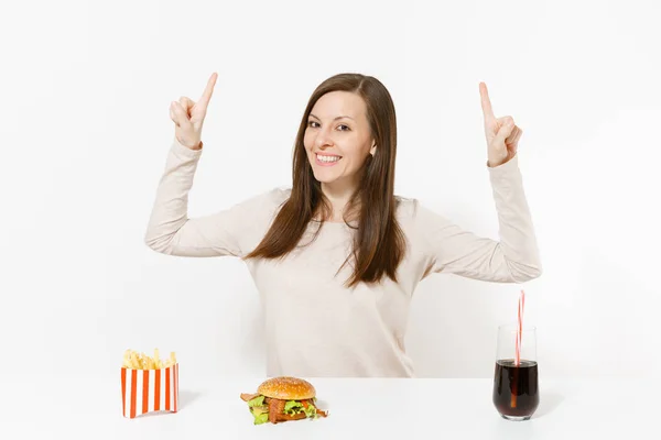 Mulher jovem apontando os dedos para cima no espaço de cópia na mesa com hambúrguer, batatas fritas, cola em garrafa de vidro isolado no fundo branco. Nutrição adequada ou fast food clássico americano. Área publicitária . — Fotografia de Stock