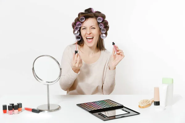 Mujer divertida con rizadores en el pelo pintando labios con lápiz labial, sentada a la mesa aplicando maquillaje con cosméticos decorativos faciales establecidos aislados sobre fondo blanco. Belleza mujer moda estilo de vida concepto . — Foto de Stock