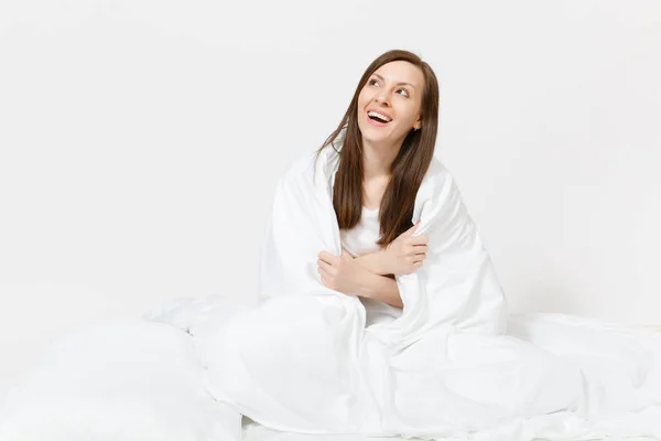 Ung brunett kvinna sitter i sängen med vitt lakan, kudde, inslagning i filt på vit bakgrund. Skönhet kvinna umgås i rummet. Vila, koppla av, gott humör koncept. Kopiera utrymme för reklam — Stockfoto