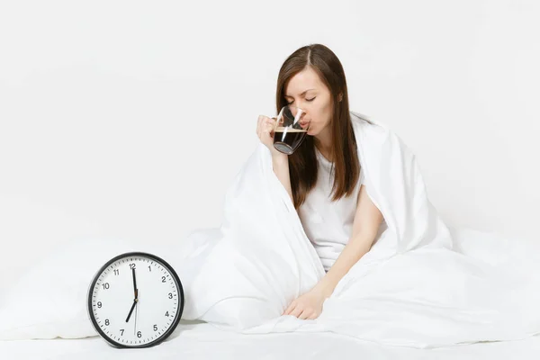 Tired sad woman sitting in bed with cup of coffee, round clock, white sheet, pillow, wrapping in blanket on white background. Female wake up early in morning, time in room. Rest, good mood concept.