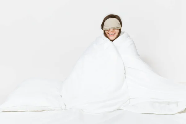 Fun tired young woman sitting in bed with sleep mask, sheet, pillow, wrapping in blanket isolated on white background. Beauty female spending time in room. Rest, relax, good mood concept. Copy space. — Stock Photo, Image