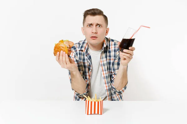 Pohledný muž znechuceně, nelidská, sedí u stolu s burger, hranolky, cola ve skle izolovaných na bílém pozadí Správná výživa nebo Klasická americká rychlé občerstvení. Reklamní plocha, kopírovat prostor. Stock Obrázky