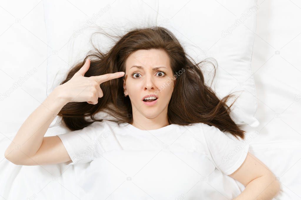Top view young woman point fingers to head as if she about shoot herself, lying in bed with white sheet, pillow, blanket. Shocked female cover ears with hand, spending time in room. Relax concept.