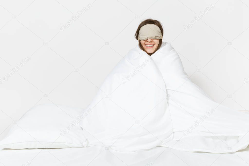 Fun tired young woman sitting in bed with sleep mask, sheet, pillow, wrapping in blanket isolated on white background. Beauty female spending time in room. Rest, relax, good mood concept. Copy space.