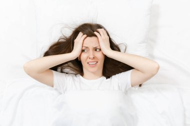 Top view of head of tired brunette young woman lying in bed with white sheet, pillow, blanket. Shocked female cover ears with hand, spending time in room. Rest, relax, good mood concept. Copy space. clipart