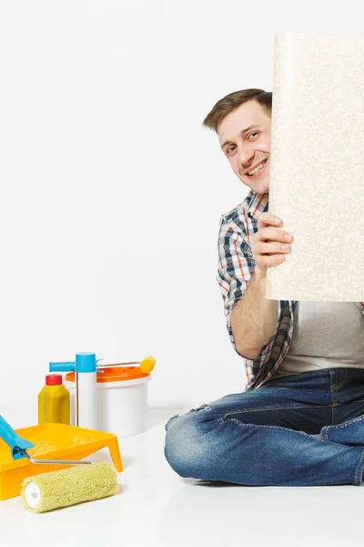 Schöner Mann sitzt auf dem Boden mit ausgeklappten Rolle von Tapeten Instrumente für die Renovierung Wohnung Zimmer isoliert auf weißem Hintergrund. Kleben Zubehör Malwerkzeuge. Reparaturheimkonzept. Kopierraum — Stockfoto