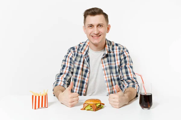 Knappe jonge man verschijnt duim, zit aan tafel met Hamburger, frietjes, cola in het glas geïsoleerd op een witte achtergrond. Goede voeding of Amerikaanse klassieke fastfood. Gebied met kopie ruimte reclame — Stockfoto
